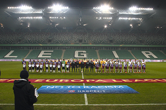 20 000 tome seter heiar på Legia. Heimelaget og gjestene frå Trabzon står oppmarsjert og klare til kamp. Kazimierz Deyna-tribunen ruvar folketom i bakgrunnen.